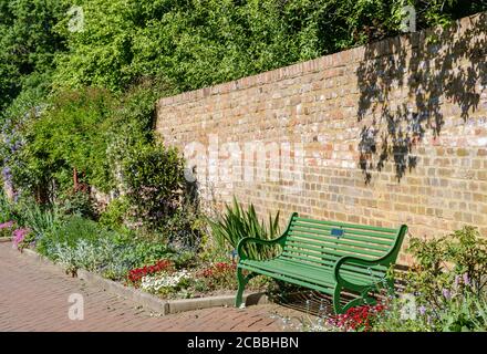 Leere grüne Parkbank mit Bäumen, Blumen und alten Backsteinmauern. Eastcote Walled Garden, Hillingdon, London Stockfoto