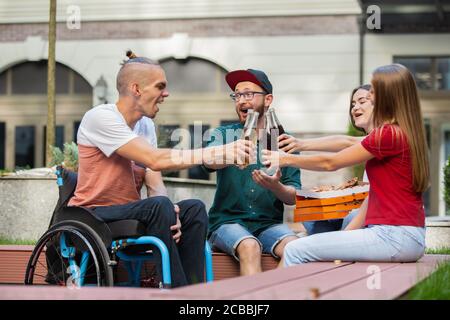 Klirren. Eine Gruppe von Freunden, die am Sommertag auf der Straße der Stadt spazieren gehen. Behinderter Mann mit seinen Freunden, die Spaß haben. Inklusion und Diversity-Konzept, normale Lebensweise von besonderen Gruppen der Gesellschaft. Stockfoto