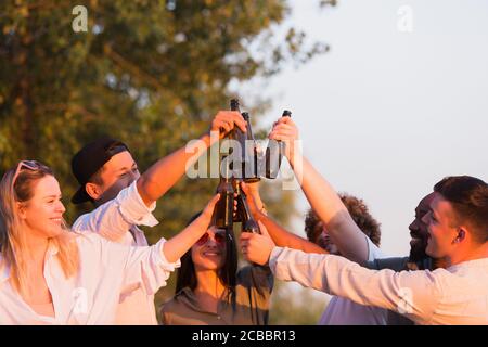 Warmes Licht. Eine Gruppe von Freunden klirren Biergläser während Picknick am Strand in der Sonne. Lifestyle, Freundschaft, Spaß haben, Wochenende und Ruhe Konzept. Sieht fröhlich, glücklich, feiern, festlich. Stockfoto