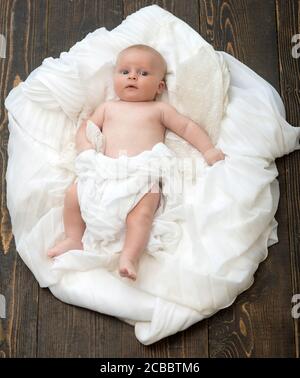 Neugeborenes Kleinkind mit blauen Augen und neugierigem Gesicht auf Holzhintergrund. Kindheit und Göttliche Geschenk Konzept. Kleinkind mit weißer Decke bedeckt. Baby liegt auf weißen weichen Bettdecke als Wolke oder Nest entworfen Stockfoto