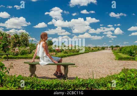 Junge Frau in weißem Kleid sitzt auf einer Bank in Rundale Palace Französisch Garten während der blühenden Saison der Rosen. Stockfoto
