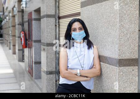 Eine Inderin mit Covid-19 Schutzmaske steht mit gekreuzten Armen in einer urbanen Unternehmensumgebung. Corporate weiblich in smart formelle Kleidung Stockfoto