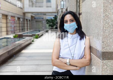 Eine Inderin mit Covid-19 Schutzmaske steht mit gekreuzten Armen in einer urbanen Unternehmensumgebung. Corporate weiblich in smart formelle Kleidung Stockfoto