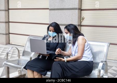 Zwei Inderinnen tragen Covid-19 Schutzmaske bei einer Diskussion auf ihrem Laptop in einem städtischen Unternehmensumgebung. Stockfoto