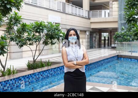 Eine Inderin mit Covid-19 Schutzmaske steht mit gekreuzten Armen in einer urbanen Unternehmensumgebung. Stockfoto