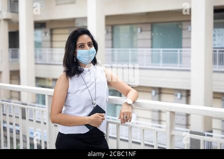 Eine Inderin mit Covid-19 Schutzmaske steht mit gekreuzten Armen in einer urbanen Unternehmensumgebung. Stockfoto
