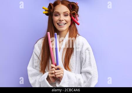 Glückliche junge Frau im Bademantel mit Lockenwicklern auf dem Haar posiert, Blick in die Kamera. Porträt, isoliert auf violettem Hintergrund Stockfoto