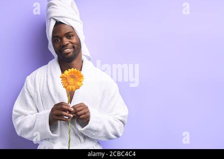 Positive schwarze Mann in Bademantel und Handtuch hält gelbe Gänseblümchen Blume isoliert über lila Hintergrund. Stockfoto