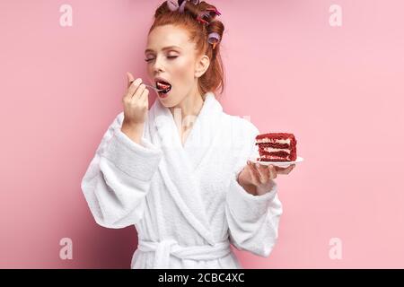 Attraktive Frau trägt Bademantel, Lockenwickler nach der Dusche essen süßen leckeren Kuchen. Emotionale Mädchen posiert mit Kuchen isoliert über rosa Hintergrund Stockfoto