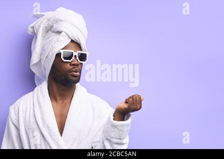 Cool schöner Mann im Bademantel und trägt Sonnenbrille tun Maniküre Für sich selbst mit Nagelfeile isoliert ober lila Hintergrund Stockfoto