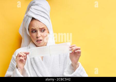Junge kaukasische Frau schauen mit Angst an war Streifen vor dem Epilieren. Tragen Bademantel und Handtuch, isoliert auf gelbem Hintergrund Stockfoto