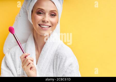 Portrait der schönen Frau in Bademantel und Handtuch hält Make-up Pinsel, Lächeln und glücklich. Kosmetikindustrie Stockfoto