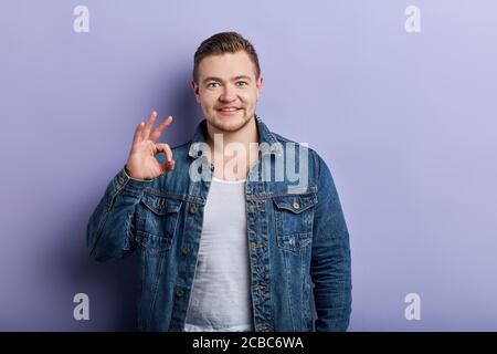 Glücklich starke muskulöse Mann in jean Jacke über blaue Wand zeigt ok Zeichen mit Fingern, Nahaufnahme Porträt, alles ist ok, okay. Stockfoto