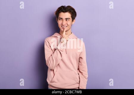Attraktive lächelnde junge asiatische Mann gekleidet lässig Blick zur Seite, berühren seine Lippen gerissen nachdenklichen Ausdruck, halten Zeigefinger auf seinem Gesicht, Stockfoto