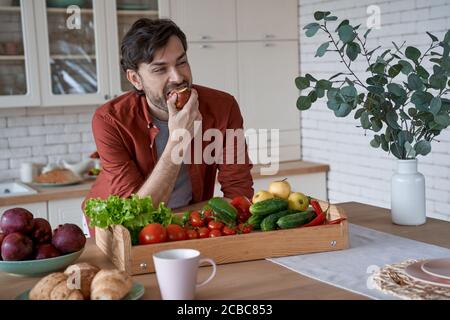 Junger glücklicher bärtiger Mann in lässiger Kleidung, der roten Apfel isst und lächelt, während er in der modernen Küche steht, Korb mit verschiedenen frischen Gemüsesorten Stockfoto