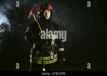 Porträt von ernst und zuversichtlich kaukasischen Feuerwehrmann Stand hält Hammer, trägt besondere Schutzuniform in der rauchigen Hintergrund Stockfoto