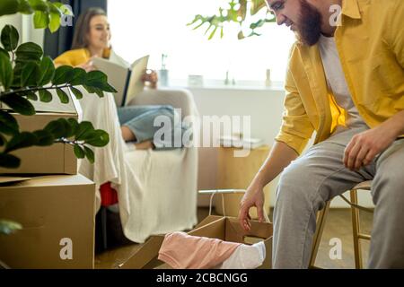 Junge kaukasische Ehepaar zu Hause, Mann Auspacken Boxen nach dem Umzug, während seine Frau liest ein Buch und spricht mit ihm. Drinnen Stockfoto