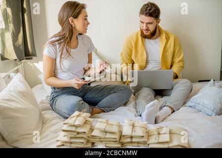 Junge kaukasische verheiratete Paar sitzen Sortierung Briefe, Einladungen zu Hause. Paar sitzen mit Laptop, sie bekommen Schlüssel aus der neuen Wohnung, senden Einladungen fo Stockfoto