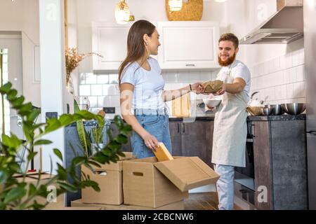 Bewegender Tag des glücklichen jungen Ehepaares Auspacken von Pappkartons, zusammen stehend in der Küche, Auspacken Karton Paket, lachen und halten Tisch Stockfoto