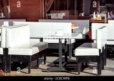 Ein weißer Holztisch mit weißen Sofas auf schwarzen Beinen Vor dem Hintergrund der Fassade mit Fenstern eines Café im Freien mit leeren Sitzen auf einem sonnigen d Stockfoto