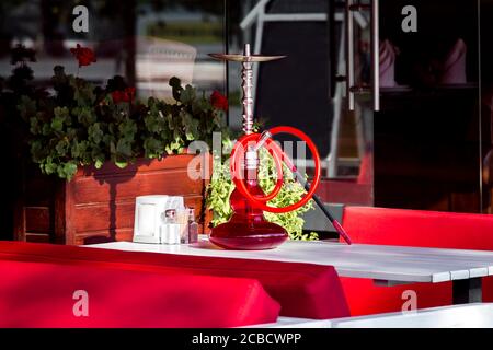 Shisha mit einem roten Glaskolben und einer Pfeife zum Rauchen auf einem weißen Holztisch eines Café im Freien mit roten Sofas an einem sonnigen Tag. Stockfoto