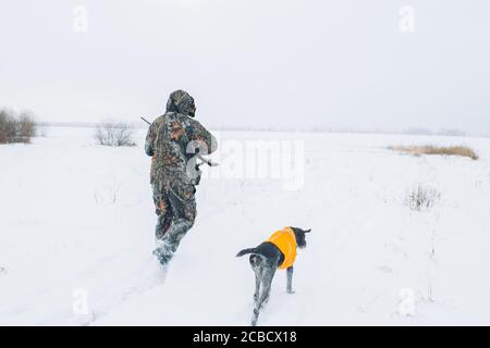 jäger und Hund zu Fuß auf dem schneebedeckten Feld. Rückansicht pghoto. Mann und sein Hund laufen nach dem getöteten Tier Stockfoto