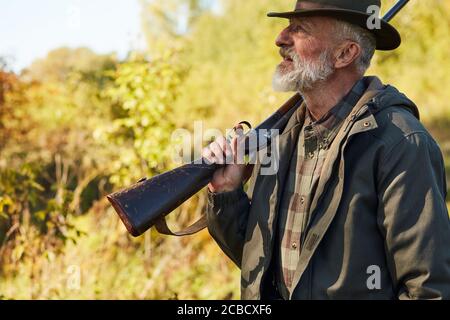 Älterer bärtiger Mann mit Waffe zur Jagd, Seitenansicht. Waldhintergrund Stockfoto