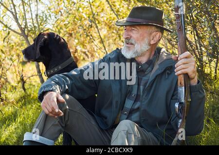 Aufmerksamer Hund und entspannender Besitzer sitzt auf Waldgras. Mann im Hut. Waffe halten. Bäume Hintergrund Stockfoto