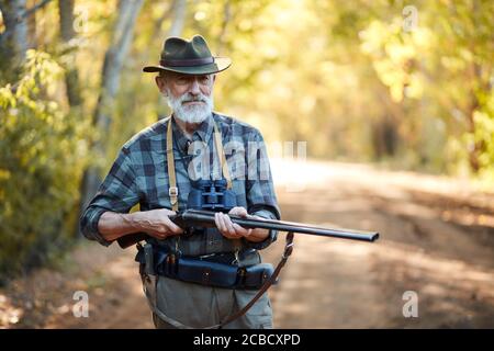 Älterer Jäger Mann mit grauem Bart halten Waffe auf Vögel jagen, bereit zu schießen. Waldhintergrund Stockfoto