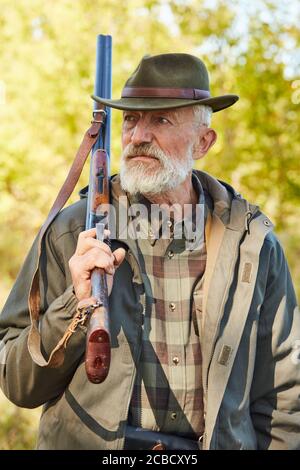Älterer kaukasischer Mann mit Jagdgewehr schaut weg, Mann mit grauem Bart trägt Jagdkleidung im Freien. Waldhintergrund Stockfoto
