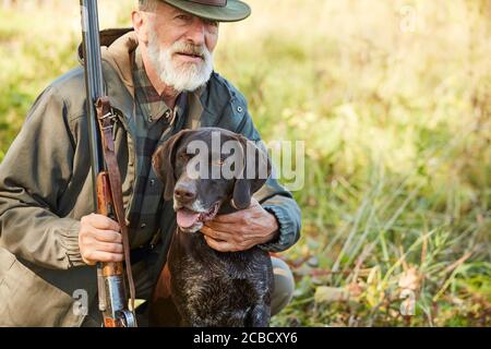 Älterer Mann umarmt seinen Jagdhund im Herbstwald. Mann trägt lässige Jagdkleidung, sitzt auf dem Boden.Waffe in den Händen Stockfoto
