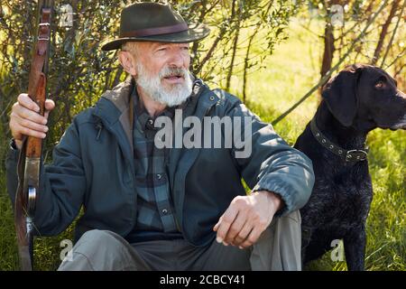 Aufmerksamer Hund und entspannender Besitzer sitzt auf Waldgras. Mann im Hut. Waffe halten. Bäume Hintergrund Stockfoto