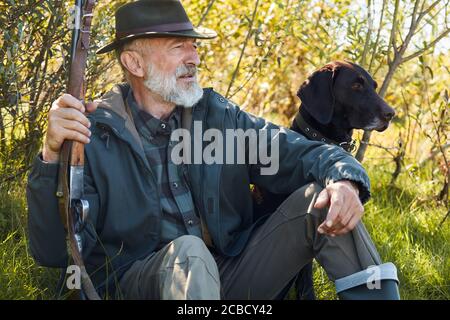 Aufmerksamer Hund und entspannender Besitzer sitzt auf Waldgras. Mann im Hut. Waffe halten. Bäume Hintergrund Stockfoto