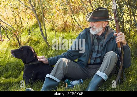 Ältere kaukasische Jäger mit seinem Gewehr, trägt Cowboy Hut sitzen auf Gras im Herbstwald. Hund sitzt nah und schaut weg Stockfoto