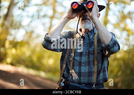 Mit Ferngläsern für bessere Jagd auf Vögel. Älterer Mann mit Fernglas während der Jagd im Herbstwald. Straßenhintergrund Stockfoto