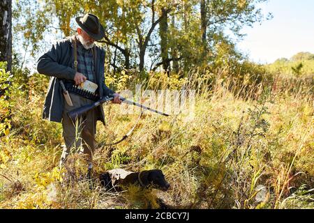 Reifer Mann auf der Jagd im Wald, gehen zu laden Gewehr, geöffnete Patrone, Hund sitzen in der Nähe von ihm Stockfoto
