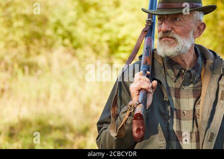 Älterer kaukasischer Mann mit Jagdgewehr schaut weg, Mann mit grauem Bart trägt Jagdkleidung im Freien. Waldhintergrund Stockfoto