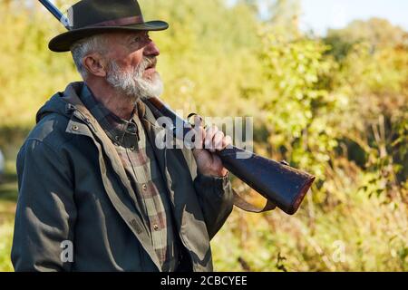 Älterer bärtiger Mann mit Waffe zur Jagd, Seitenansicht. Waldhintergrund Stockfoto