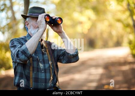 Mit Ferngläsern für bessere Jagd auf Vögel. Älterer Mann mit Fernglas während der Jagd im Herbstwald. Straßenhintergrund Stockfoto