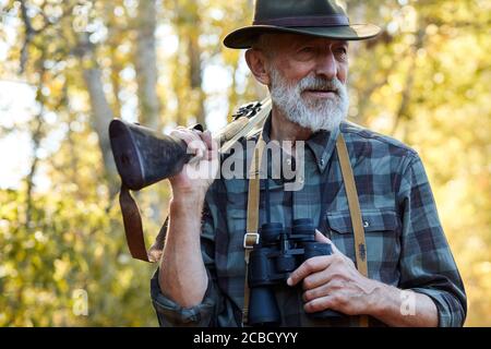 Zufriedener Mann mit grauem Bart schaut weg, steht mit Fernglas in einer Hand, Schrotflinte auf den Schultern. Wald, Herbstbäume Hintergrund, sonniger Tag Stockfoto