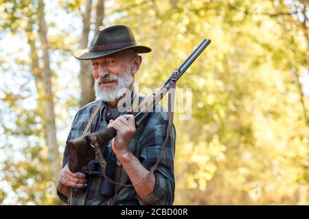 Happy Senior Hunter Lächeln nach immer gut, große Trophäe im Wald. Jagd auf wilde Tiere, Vögel. Zufriedener Jäger mit grauem Bart, mit Schrotflinte Stockfoto