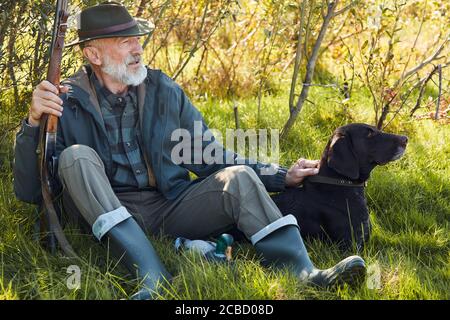 Ältere kaukasische Jäger mit seinem Gewehr, trägt Cowboy Hut sitzen auf Gras im Herbstwald. Hund sitzt nahe, beide schauen weg Stockfoto