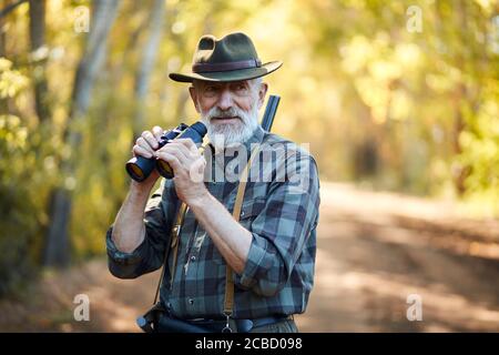 Mit Ferngläsern für bessere Jagd auf Vögel. Senior Mann hält Fernglas während der Jagd im Herbst Wald Blick auf Kamera. Straßenhintergrund Stockfoto