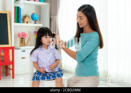 Asiatische Mutter kämmt die Haare ihrer Tochter am Morgen, bevor sie zur Schule im Wohnzimmer zu Hause gehen. Mutter Morgen tägliche Routine-Konzept. Stockfoto