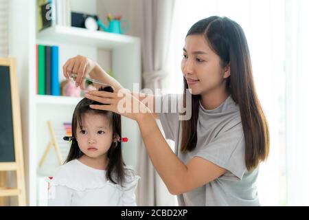 Asiatische Mutter Haare schneiden ihre Tochter im Wohnzimmer zu Hause während zu Hause bleiben sicher von Covid-19 Coronavirus während der Sperrung. Selbstquarantäne an Stockfoto