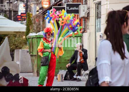 Bukarest / Rumänien - 25. August 2019: Frau als Clown verkleidet, die Luftballons auf der Straße verkauft und Mann in Anzug, der mit Geige musiziert Stockfoto