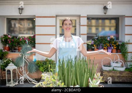 Hübsche lächelnde Frau in Schürze, die vor dem Tisch mit Blumen steht und sie zeigt Stockfoto