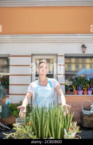 Hübsche lächelnde Frau in Schürze, die vor dem Tisch mit Blumen steht und sie stolz zeigt Stockfoto