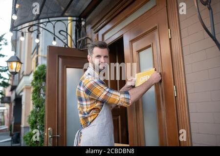 Junger bärtiger Kellner in grauer Schürze, der ein Schild an der Tür aufhing und lächelte Stockfoto