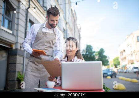 Braunhaarige Frau, die am Tisch sitzt, das Menü hält und sich mit dem bärtigen Kellner berät Stockfoto